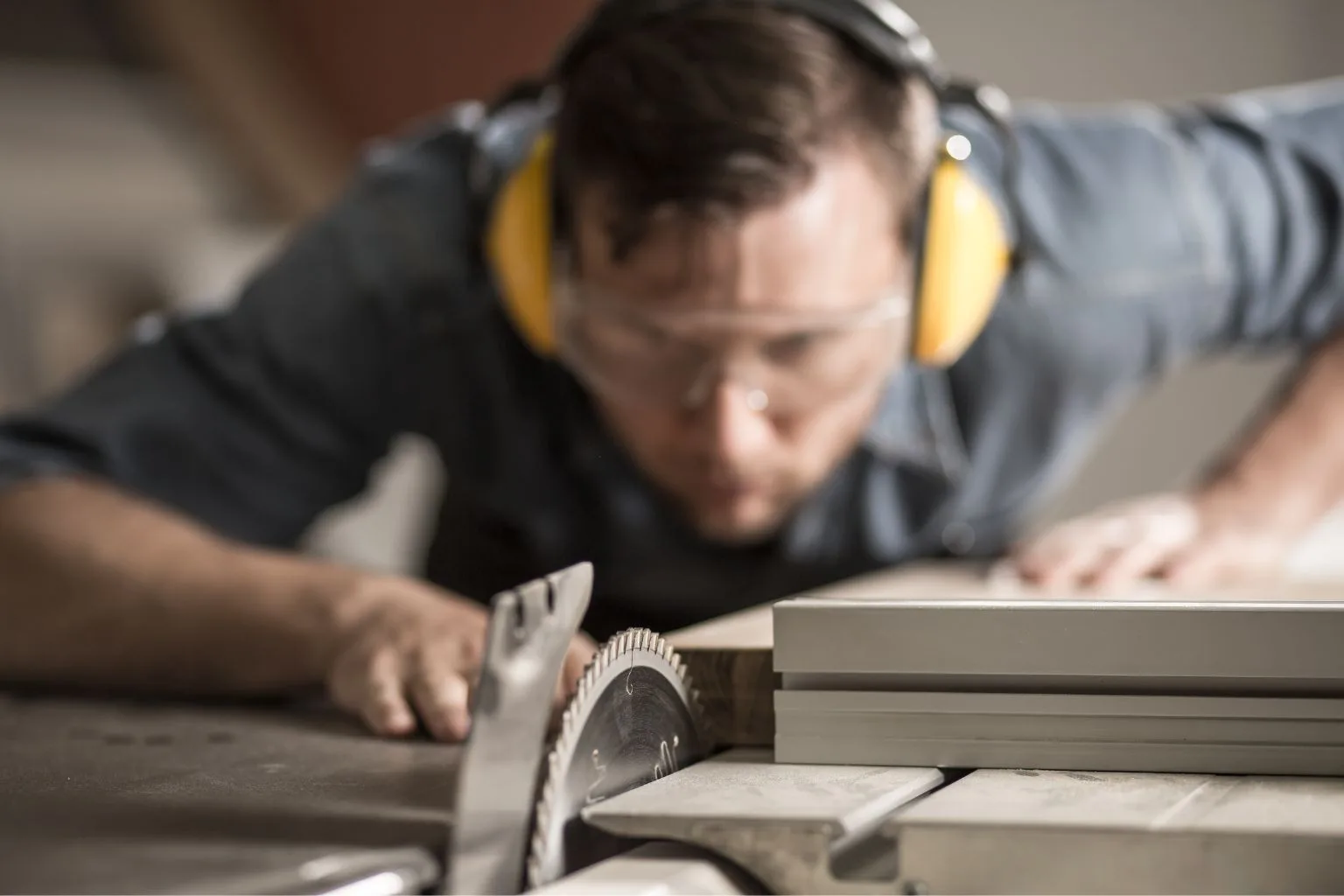 A woodworker wearing safety glasses and earmuffs carefully aligns a piece of wood on a table saw, focusing intently on the blade. The image highlights the importance of precision and safety in woodworking